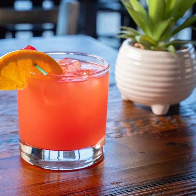 Bright coral cocktail with orange garnish on a wooden table with a small plant nearby.