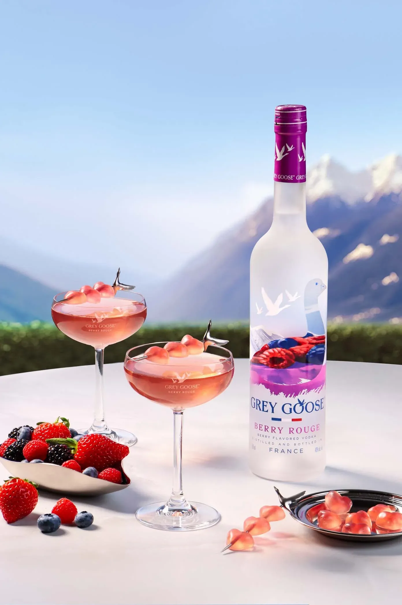A bottle of Grey Goose Berry Rouge vodka on a table with two pink cocktails garnished with fruit, a bowl of mixed berries, and a mountain landscape in the background.