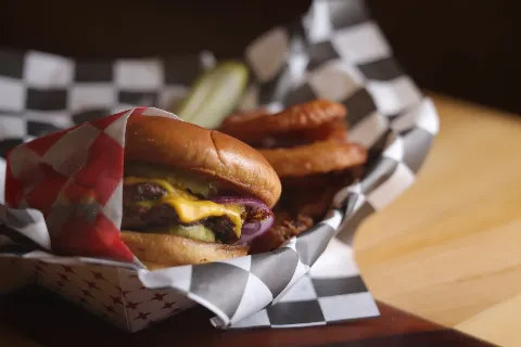 Close-up of a smash burger with pickles, onions, and melted cheese, served in a checkered paper basket with a side of onion rings.