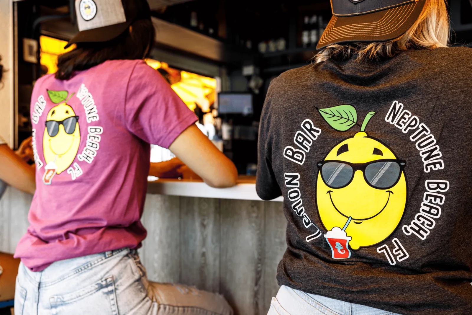 Two people wearing t-shirts with a lemon character wearing sunglasses and sipping a drink at a bar.