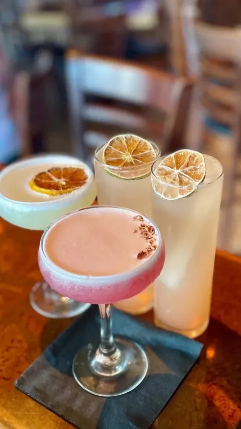 Four colorful cocktails on a dark napkin on a wooden table, with blurred chairs and bar in the background. Two drinks are in tall glasses with lemon or lime slices, one is in a stemmed glass with a pi
