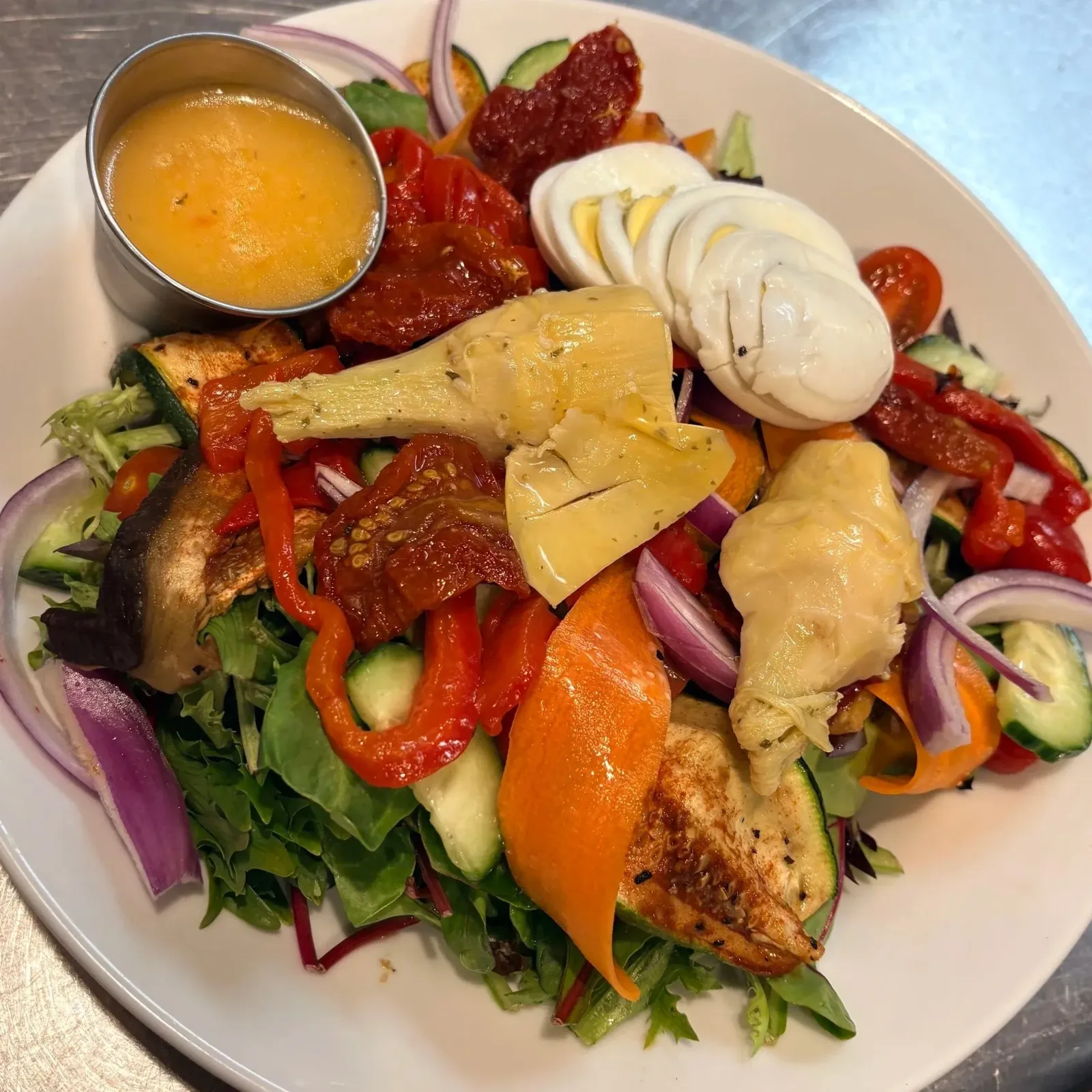 Mixed salad with grilled chicken, sliced boiled eggs, cherry tomatoes, red onions, cucumber, roasted red peppers, artichoke hearts, and a small container of yellow dressing.