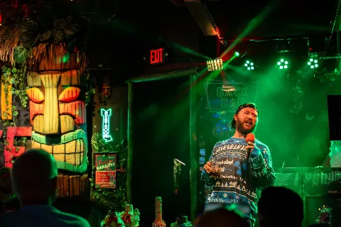 A man with a beard and a backwards cap holding a microphone presents on a stage with colorful lighting and tropical decor, including a large tiki face and neon signs, in a nightclub or bar setting.