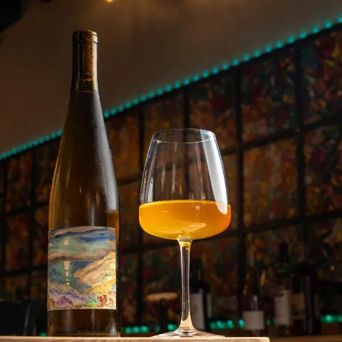 A bottle of skin contact wine and a half full glass sit on a bar counter.