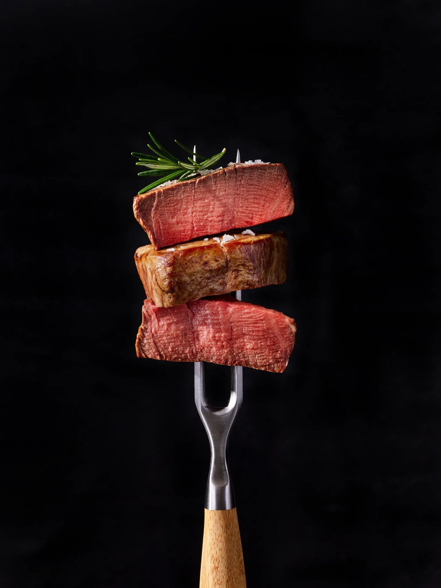 Three slices of medium-rare steak stacked on a fork with a sprig of rosemary on top against a black background.