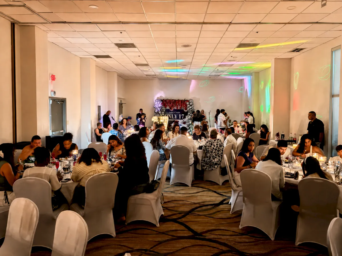 The image shows a decorated event hall with people seated at round tables, likely attending a social or formal gathering.