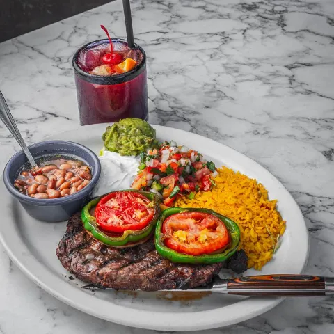 Grilled steak topped with peppers, served alongside rice, beans, salsa, and cocktail.