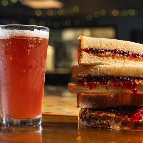 Fruit beer beside stacked peanut butter and jelly sandwiches on a counter.