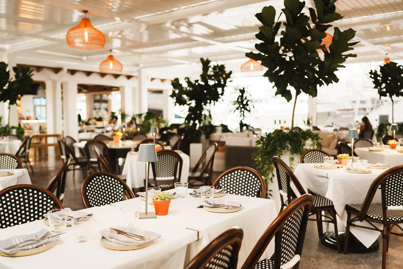 Sophisticated dining room at Lamarina waterfront restaurant in West Palm Beach featuring elegant table settings, woven chairs, pendant lighting, and lush greenery for an elevated coastal dining experi