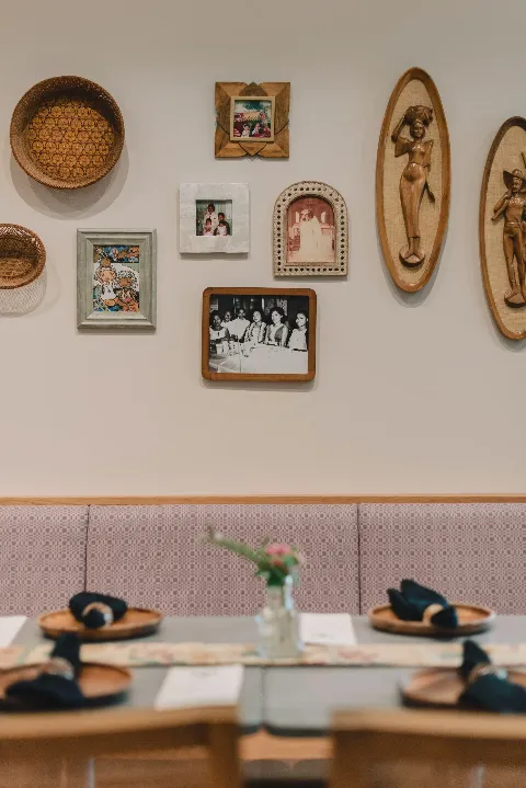 Dining area with decorative wall art, framed photos, and wooden carvings; table with napkins and a flower vase.