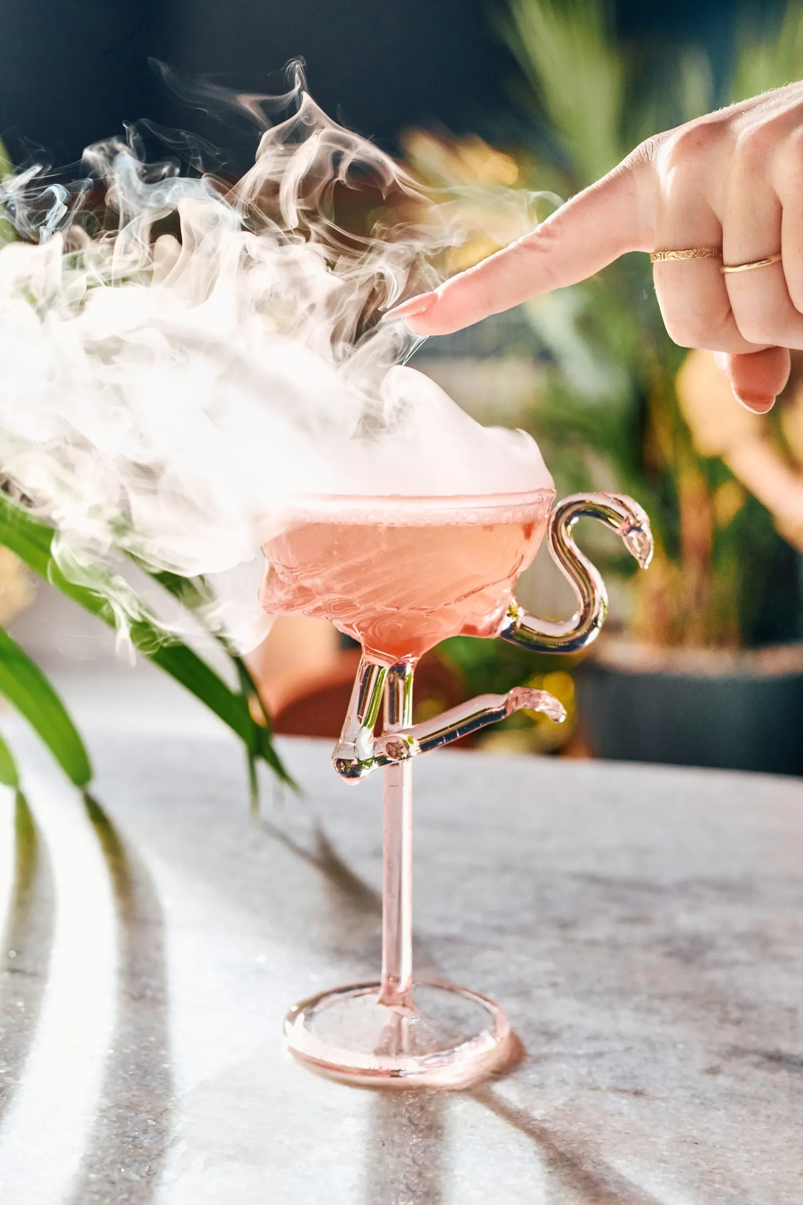 A close-up of a hand with a gold ring touching the top of a shimmery pink cocktail in a decorative glass with a flaming effect and smoke rising from it. The background is blurred with green foliage.