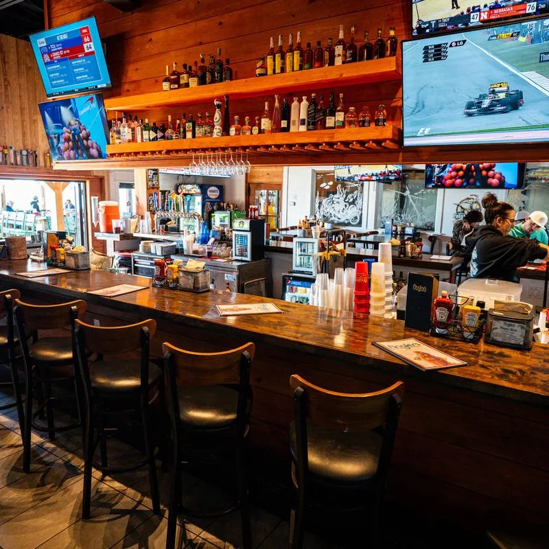 Bar area with a counter, stools, shelves with bottles and TV screens on the wall.