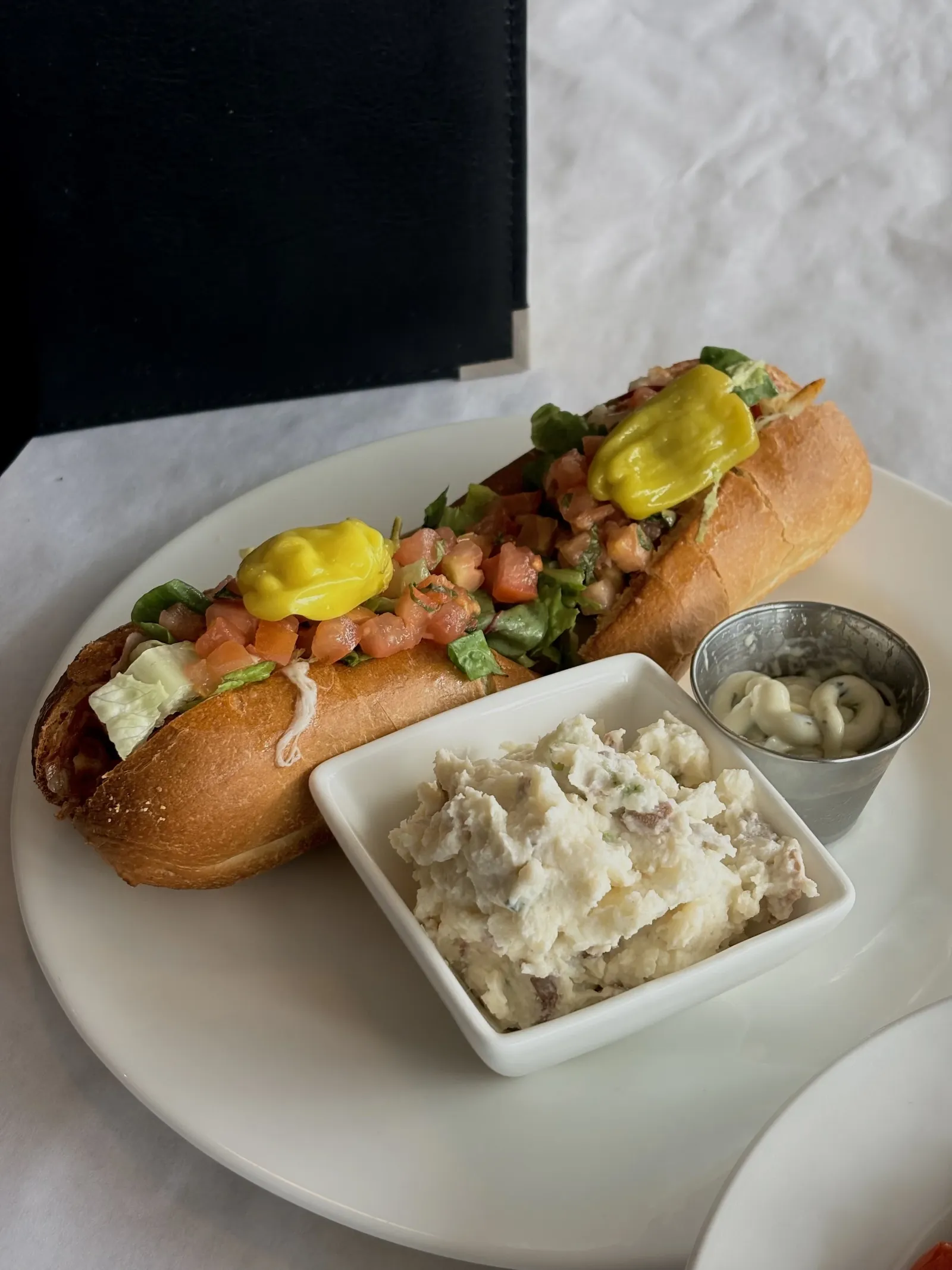 Hot dog with relish and chopped vegetables, served with potato salad and a small cup of mayonnaise or tartar sauce on a white plate.