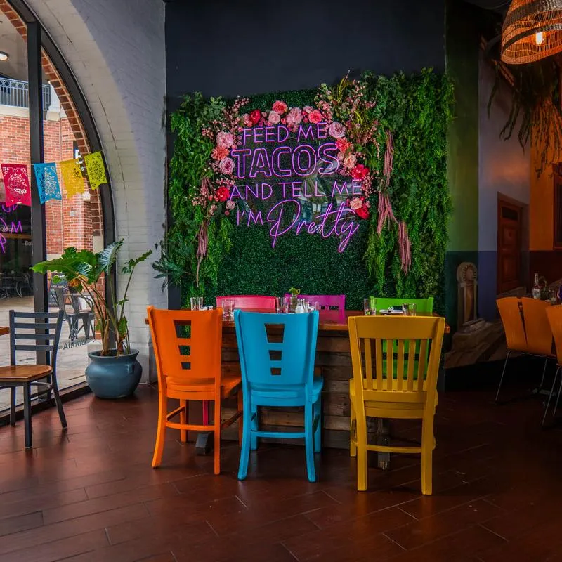 Interior with vibrant greenery decor and a neon sign, tables and colorful chairs.