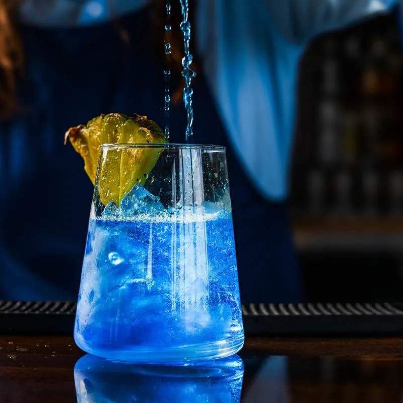 Blue cocktail being strained into a glass garnished with a fresh pineapple wedge.