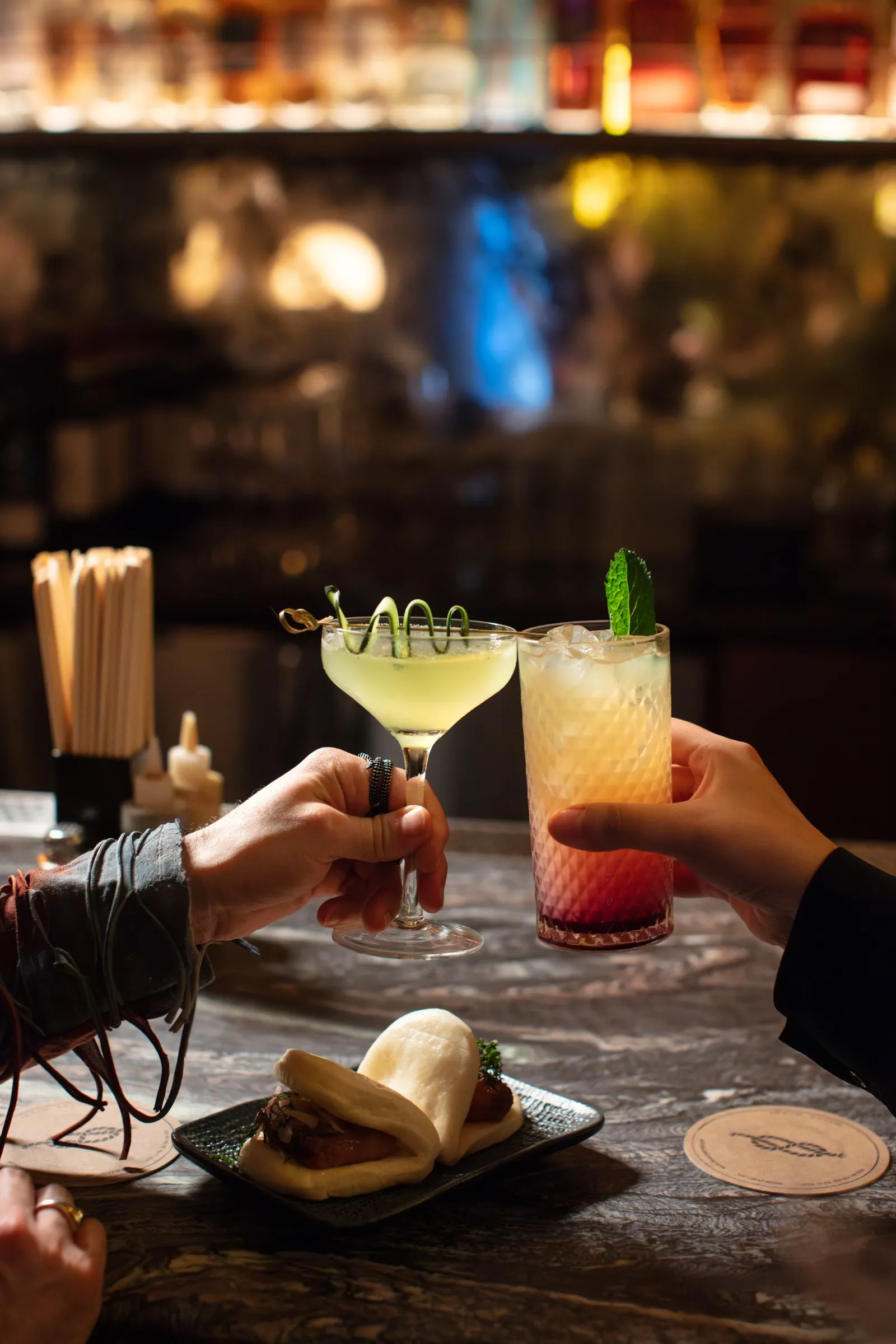 Two people toasting with cocktails at a bar, with a plate of hot dogs and buns on the table, in a cozy, dimly-lit restaurant setting.