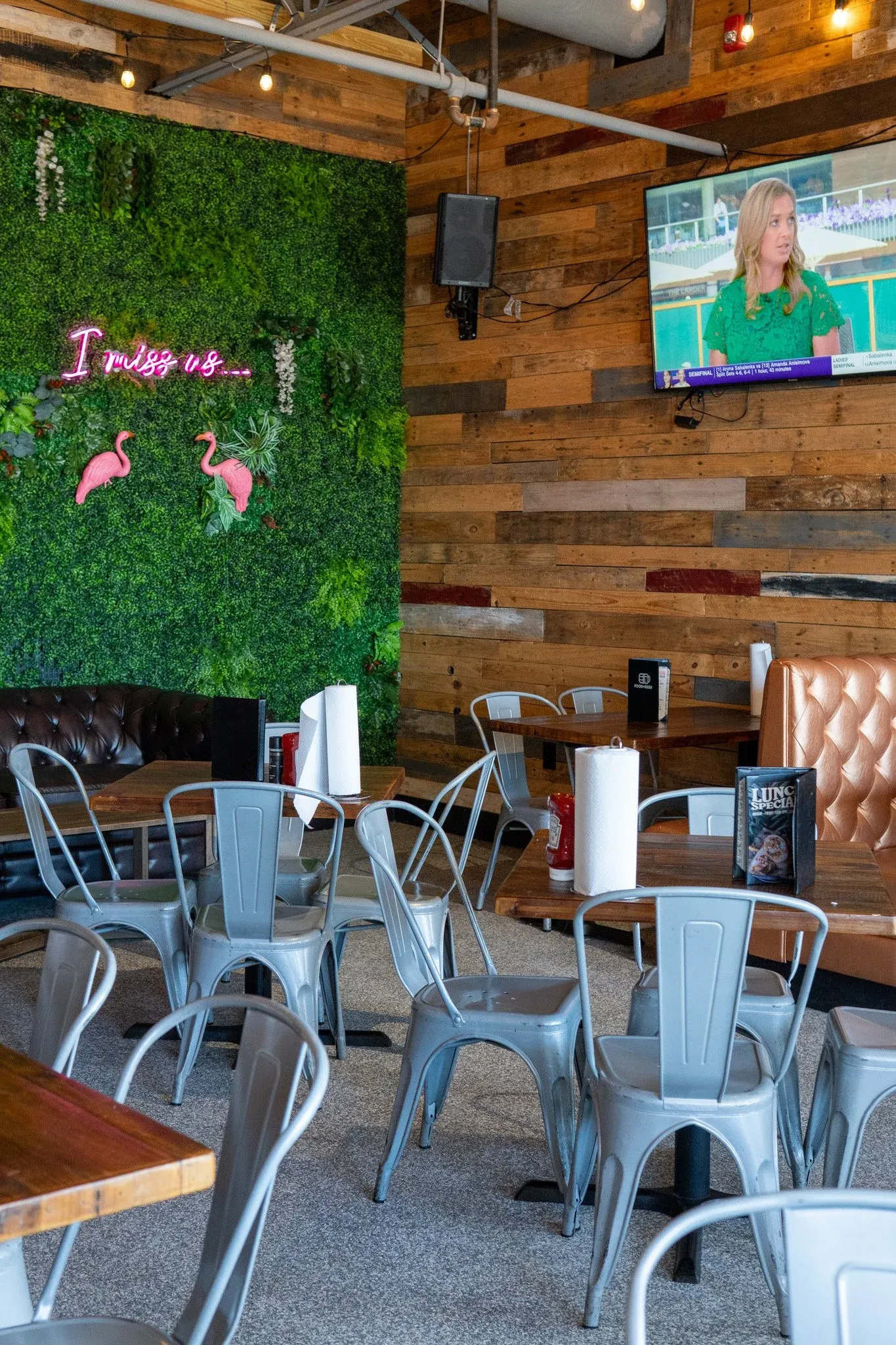 Interior of a casual restaurant with wooden walls, a green foliage wall with a pink neon sign that says 'I miss us...' and flamingo decorations. There is a flat-screen TV mounted on the wall, metal ch