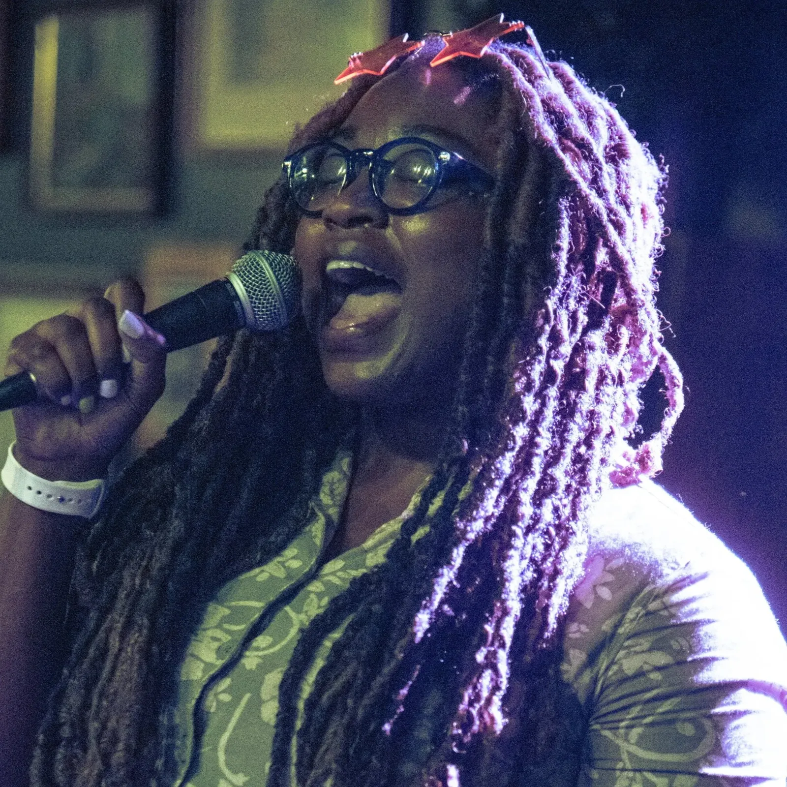 A woman with purple Dreadlocks wearing glasses and a white wristband, singing into a microphone.