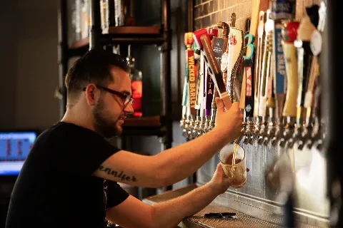 A bartender pours beer from a tap into a glass at a bar with multiple beer taps lined up.