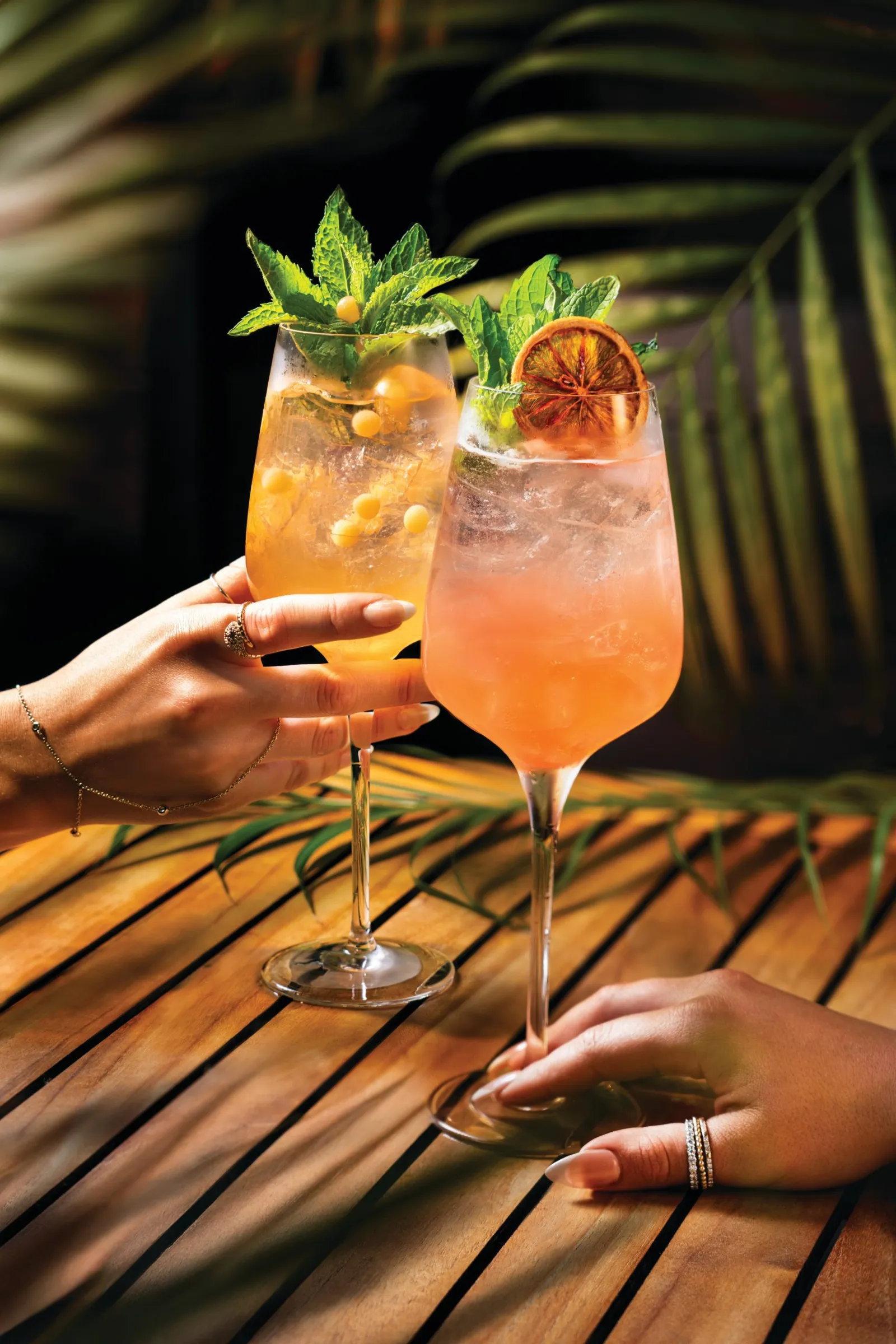 Two hands holding large wine glasses filled with colorful cocktails garnished with mint, berries, and citrus, set on a wooden table with tropical leaves in the background.