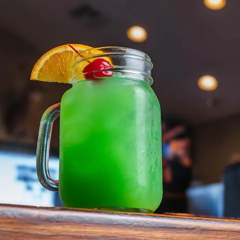 Green cocktail with orange and cherry garnish on a counter.
