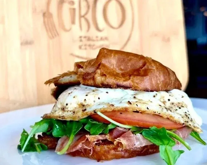 Close-up of a breakfast sandwich with a fried egg, tomato, greens, ham, and a toasted brioche bun, on a white plate.