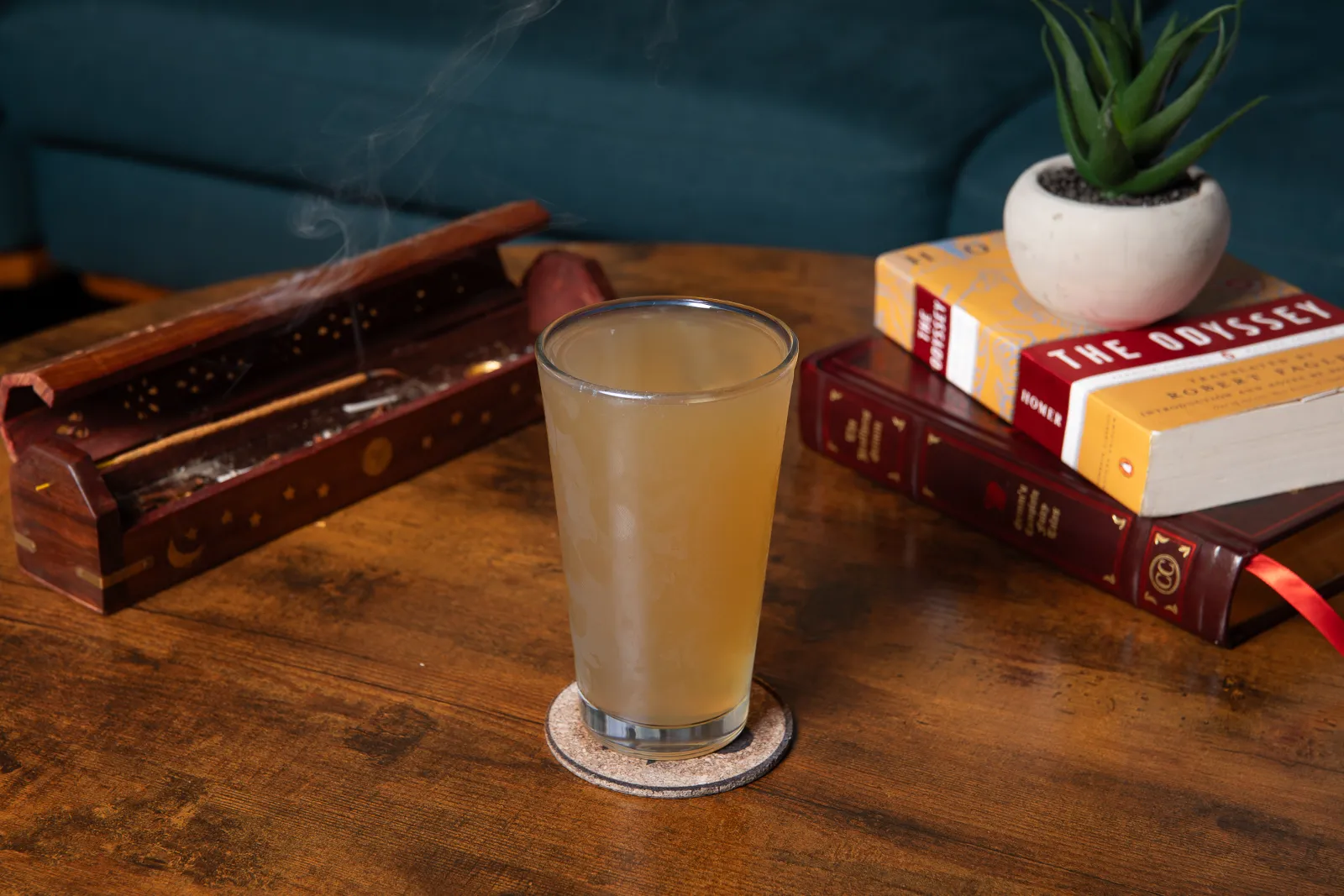 A glass of ice-cold euphoric tea on a coaster, on a wooden table, with a box of matches, a small potted succulent plant, and a stack of books including 'The Odyssey' and a red leather-bound book.