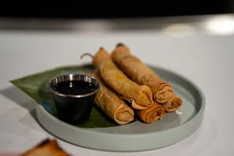 Three spring rolls on a green leaf on a white plate with a small cup of dark soy sauce