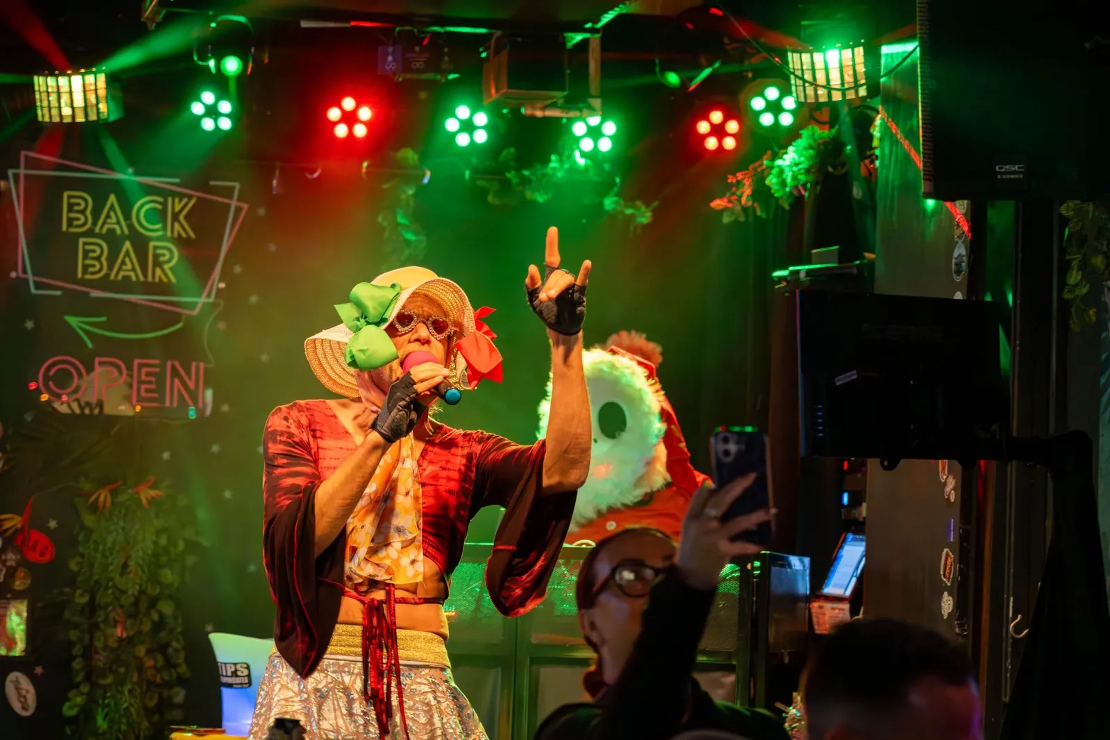Performer with a wide-brimmed straw hat decorated with colorful bows, sunglasses, and a microphone on stage in a bar with neon signs, colorful lights, and someone taking a picture.