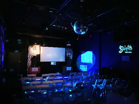 Dark room with chairs arranged for an event, screen, disco ball, and neon sign.