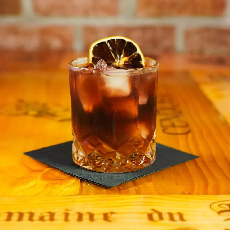 Dark cocktail over ice garnished with dry citrus, served on a wooden table.