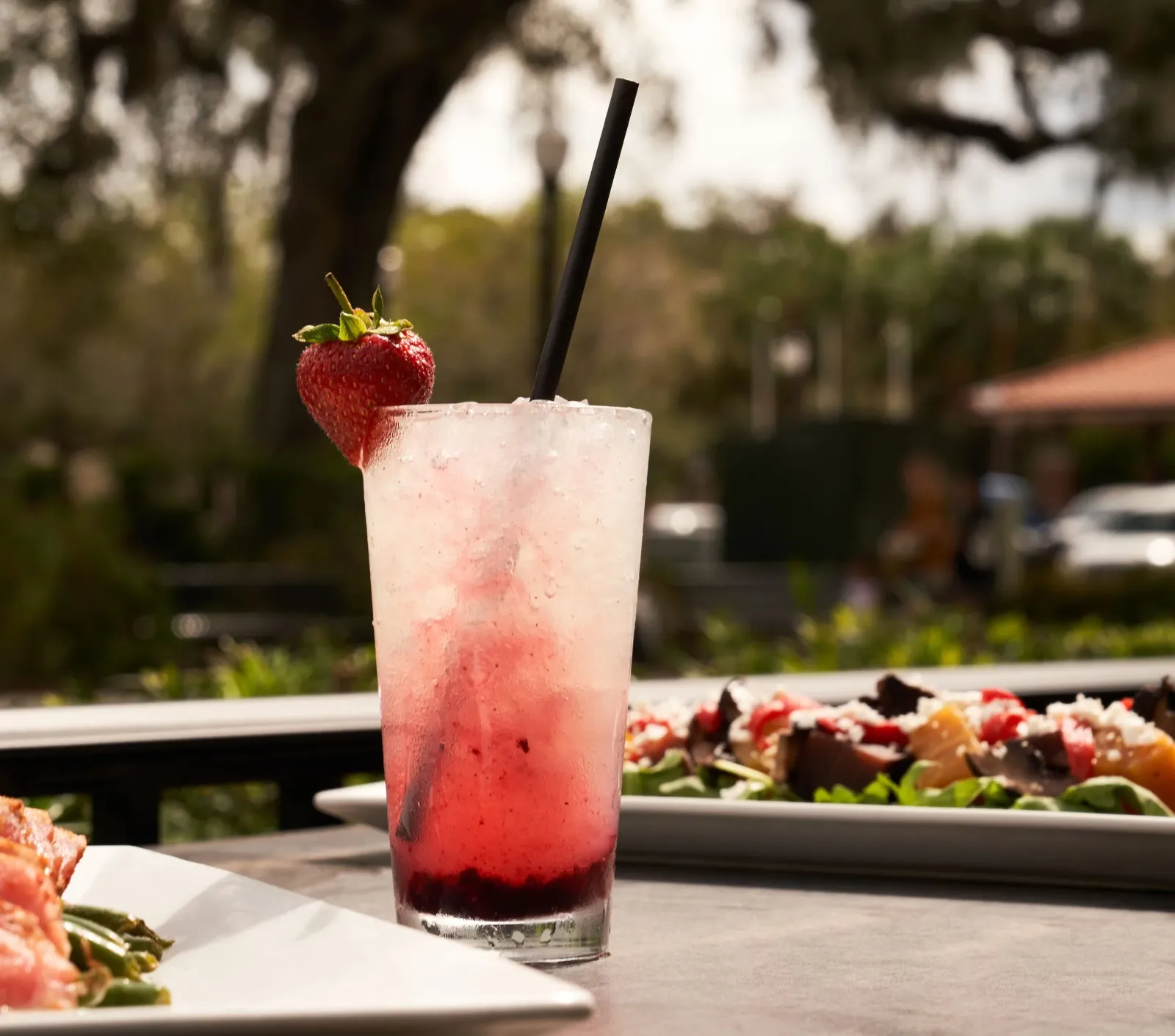 A tall glass of strawberry cocktail with ice and a strawberry garnish and straw, on a table outdoors with plates of food in the background.