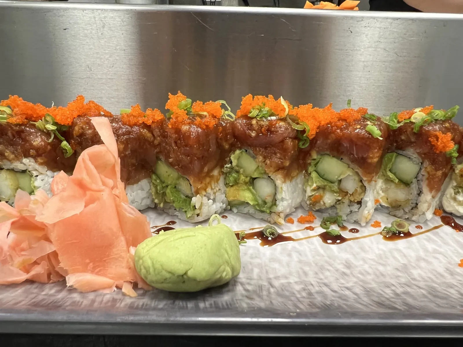 A sushi roll topped with fish roe, drizzled sauce, and chopped green onions, served on a metallic tray with slices of pickled ginger and a small mound of wasabi.