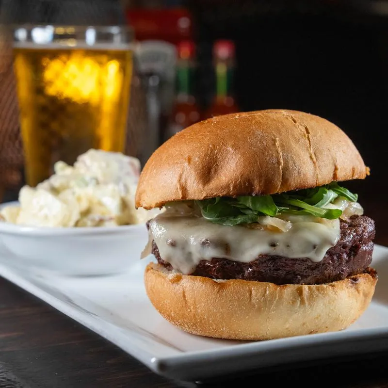 Gourmet burger topped with melted cheese and arugula, accompanied by a potato salad.