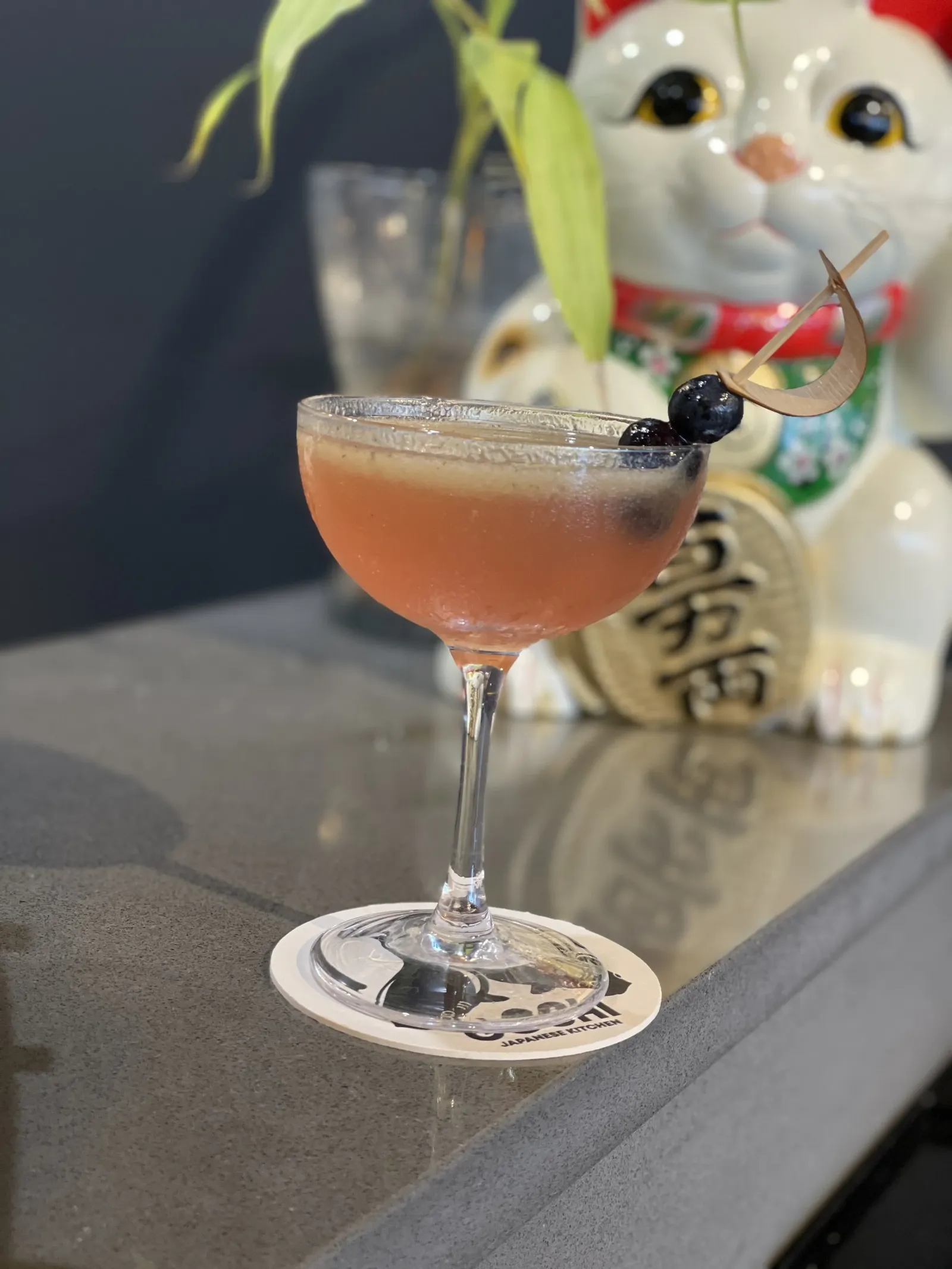 A pink cocktail with blueberries and a decorative cherry on top in a coupe glass, with a Japanese lucky cat figurine and a potted plant in the background.