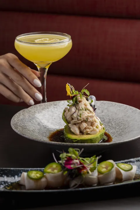Yellow cocktail in a coupe glass served with crab avocado salad and yellowtail crudo