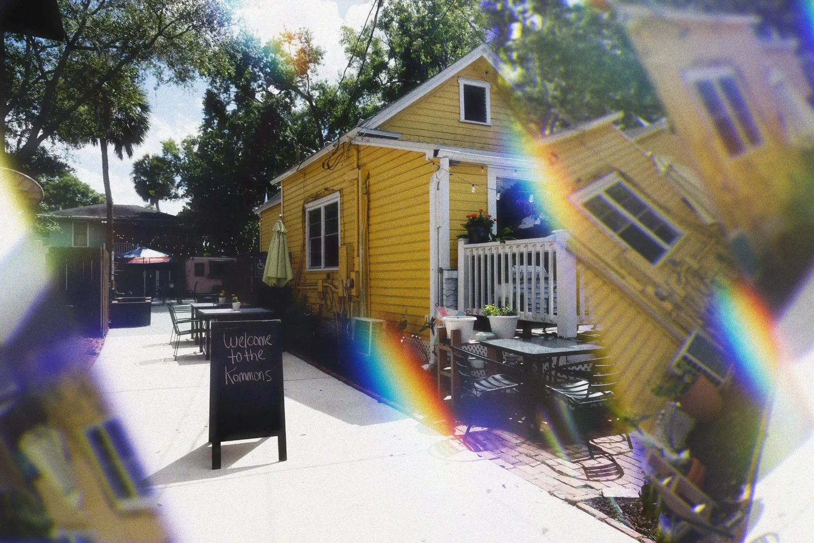 Kaleidoscope Thornton Park, a yellow building with white trim and a small porch, outdoor patio with tables and chairs, potted plants, sign that says 'Welcome to the Kommons', trees and partly cloudy s