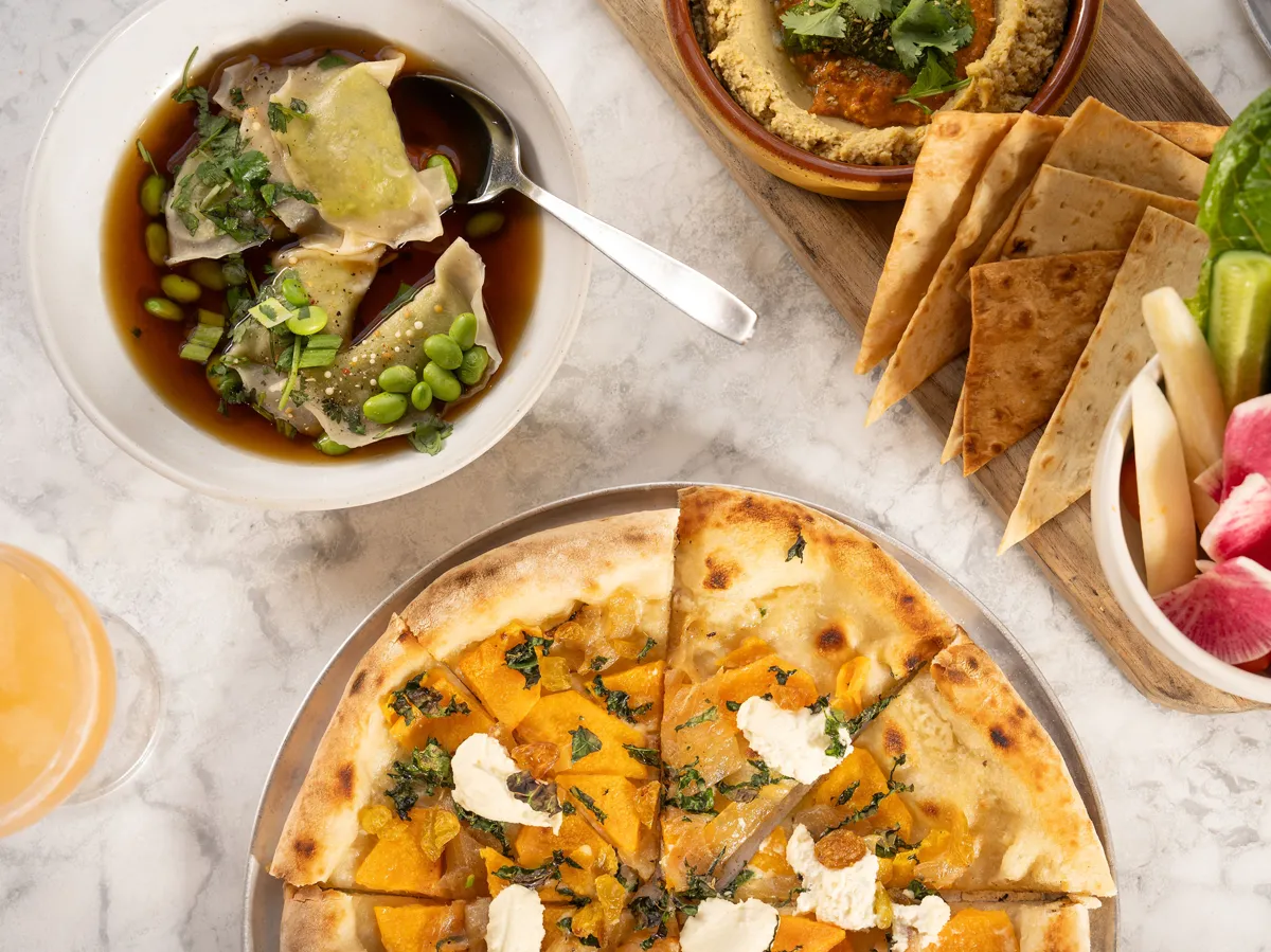 A variety of dishes: dumplings, pizza, hummus with pita, and vegetables, presented on a marble surface with a drink.