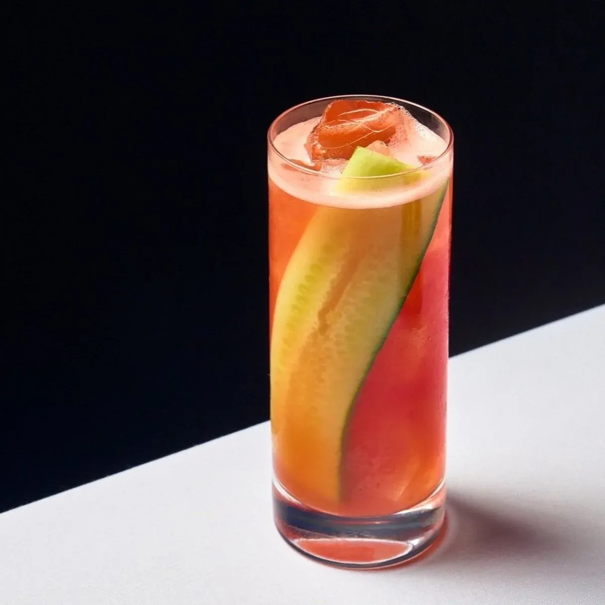Tall glass with pink cocktail, cucumber slice, ice, and rose-shaped garnish on black background.