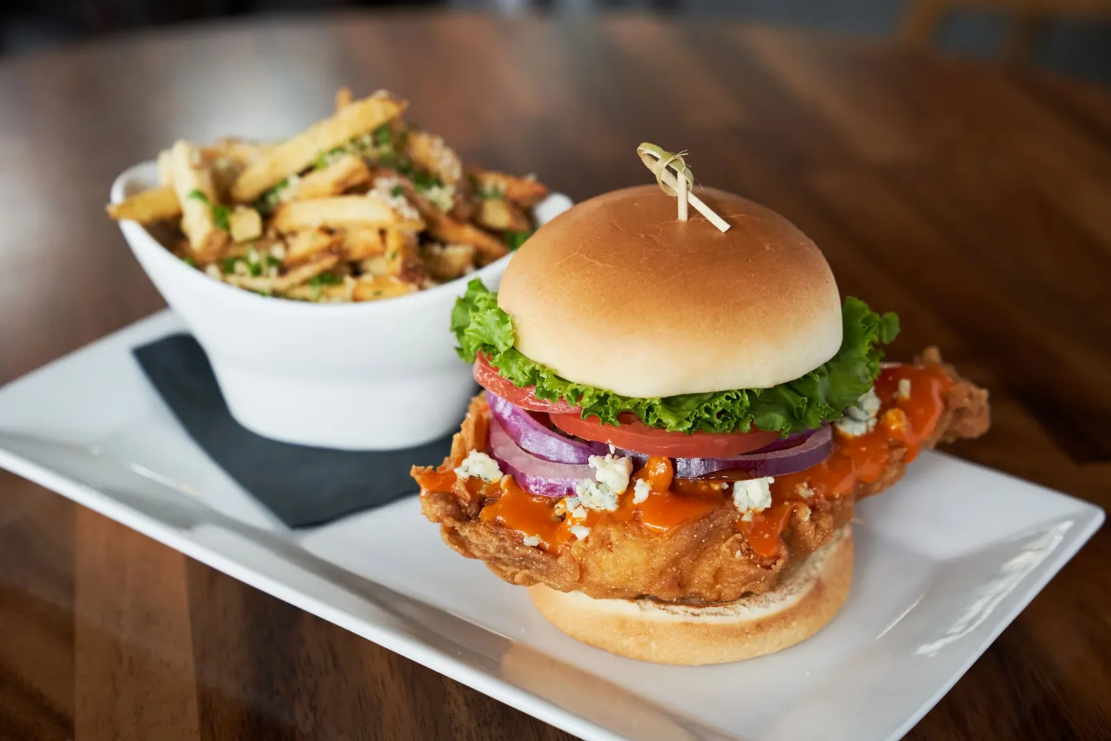 Fried chicken sandwich with lettuce, tomato, onion, blue cheese, and hot sauce on a bun, served with a side of seasoned French fries.