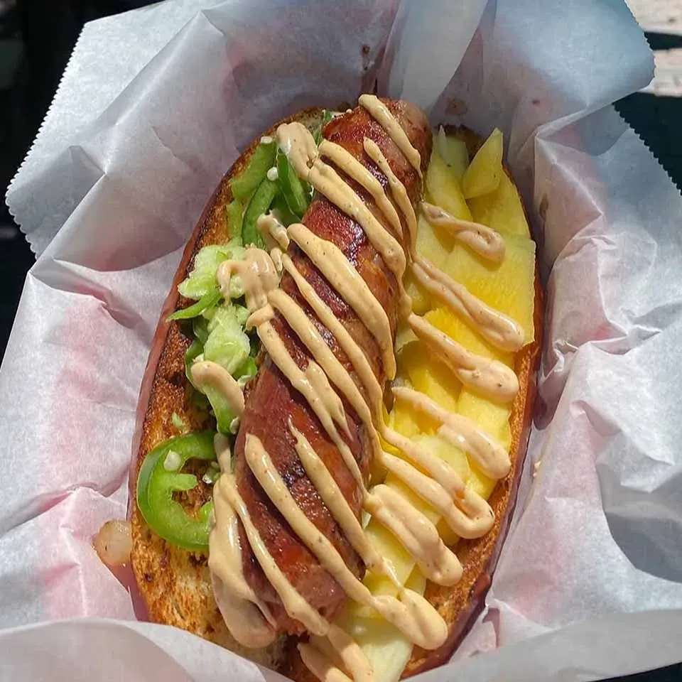 A bacon-wrapped hot dog with jalapeños, pineapple, and creamy sauce in a toasted bun.