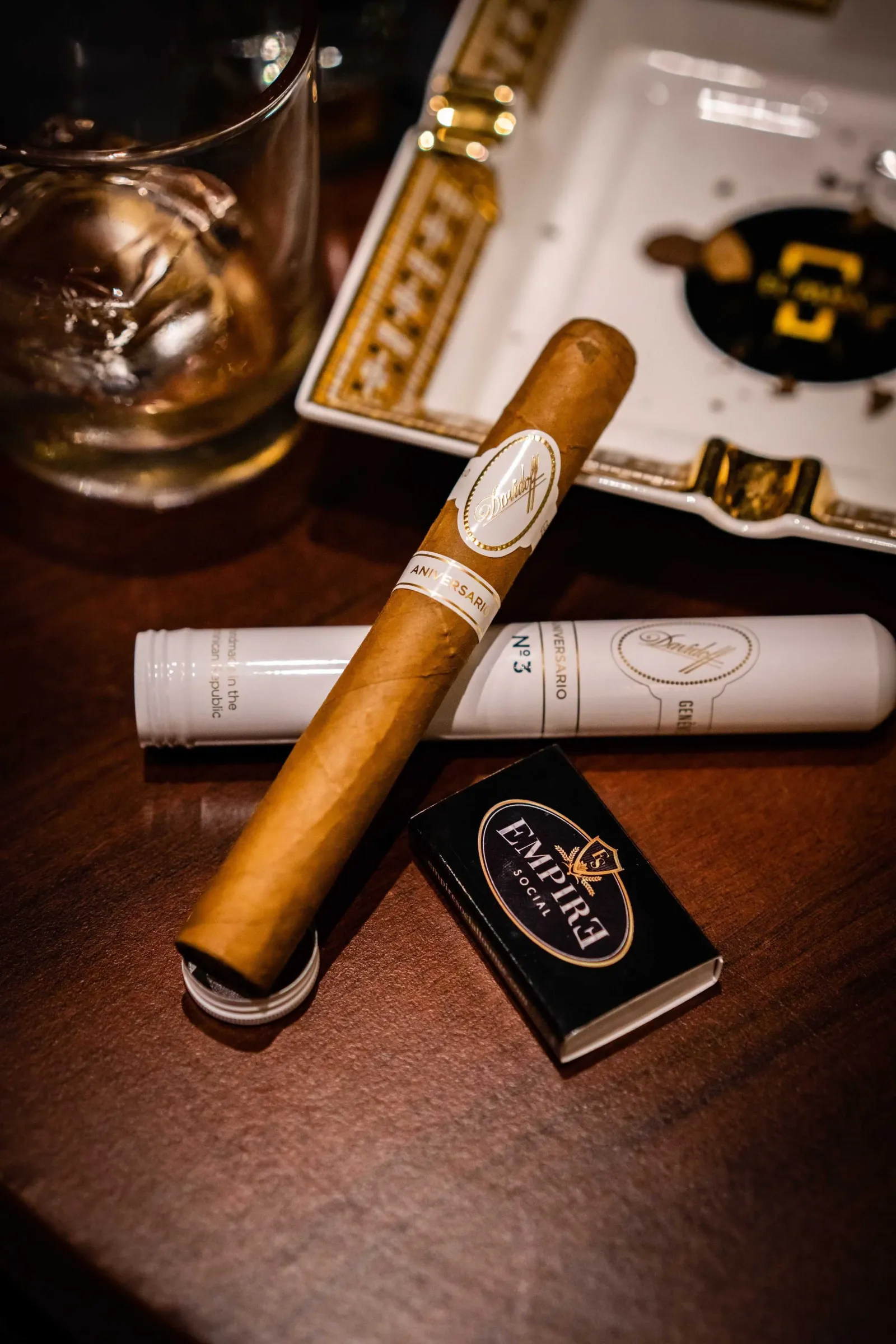 A cigar, a glass of whiskey with ice, a box of cigars, and a pack of Eclipse Social chewing gum on a wooden table.