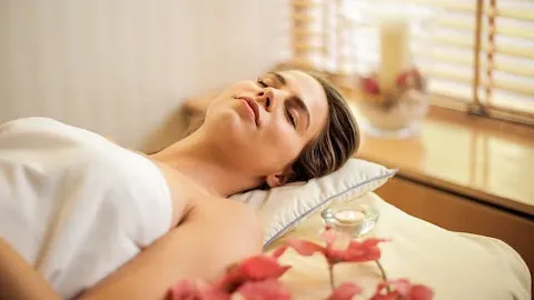 A person is relaxing on a massage table surrounded by flowers and soft lighting, suggesting a serene spa environment.