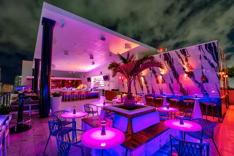 Rooftop bar at night with pink‑purple lighting, round pink tables, metal chairs, and a central planter.