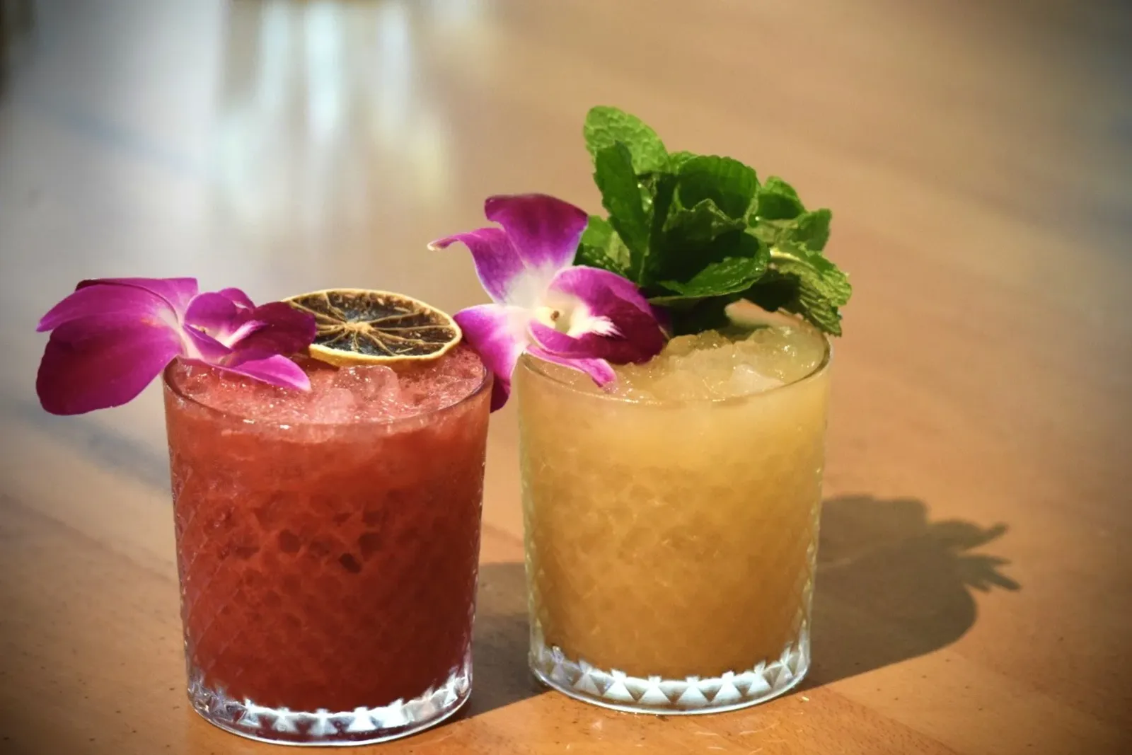 Two colorful cocktails in textured glasses garnished with edible flowers, lemon slices, mint leaves, and crushed ice on a wooden surface.