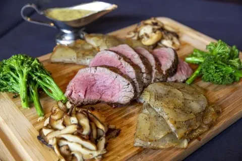 Sliced beef tenderloin with vegetables, mushroom, and sauce on a wooden platter.