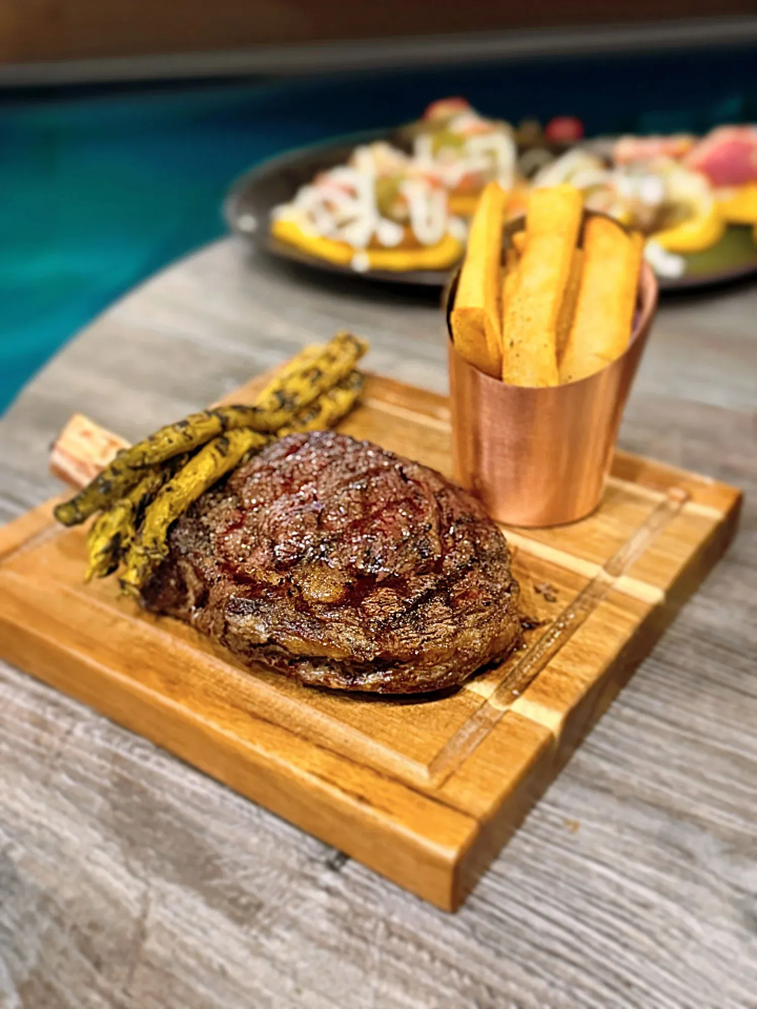 Grilled steak on a wooden board with asparagus and a copper cup of fries.