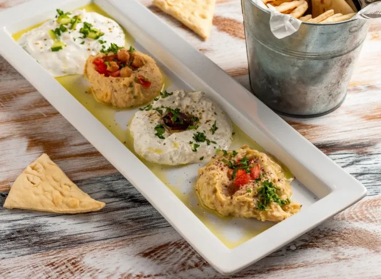 Greek cuisine spread at Acropolis Taverna, showcasing the Appetizer Sampler which comes with Hummus, Tirosalata, Tzatziki, and Baba Ganouj. Served with fresh pita.