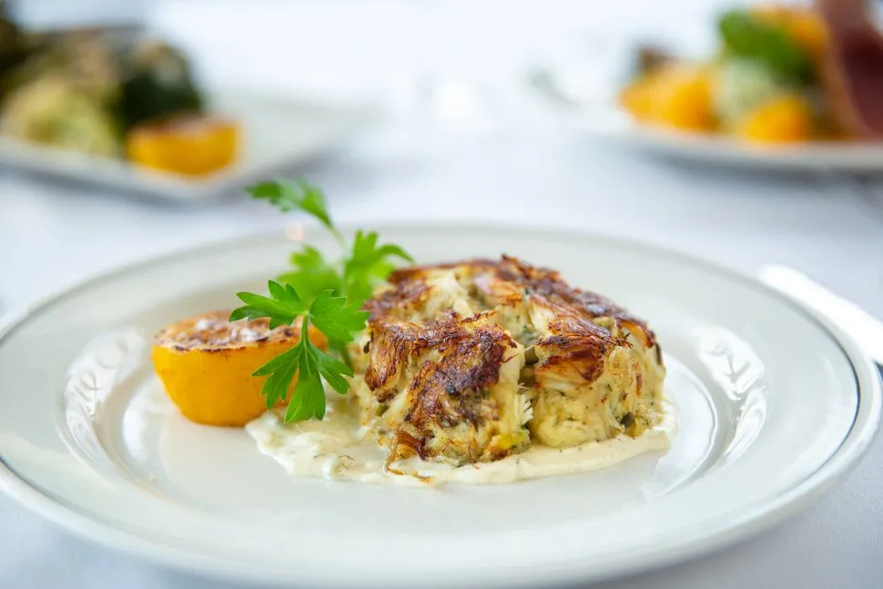 Jumbo lump crab cake with a golden-brown crust, served over a light cream sauce and garnished with fresh parsley and a roasted lemon half on a white plate.