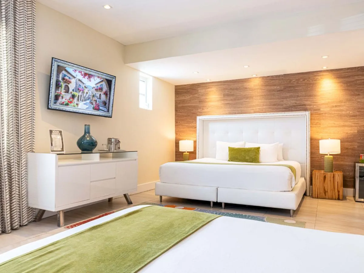 A modern hotel-style bedroom with a white upholstered bed, wooden feature wall, two nightstands, a dresser, and a wall-mounted TV.